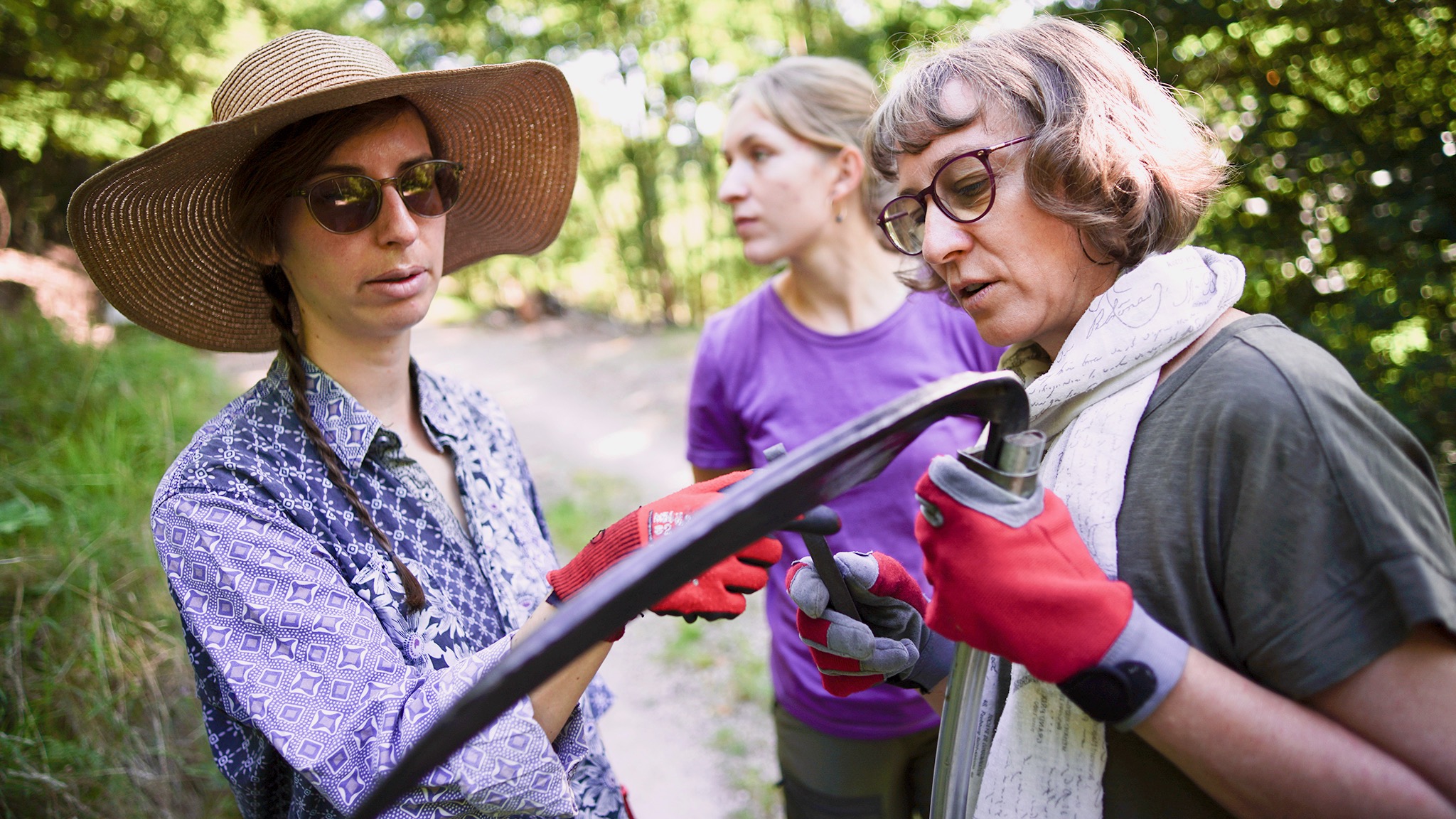Eine Frau mit großen Strohhut und Sonnenbrille betrachtet mit einer anderen Frau ein Sensenblatt