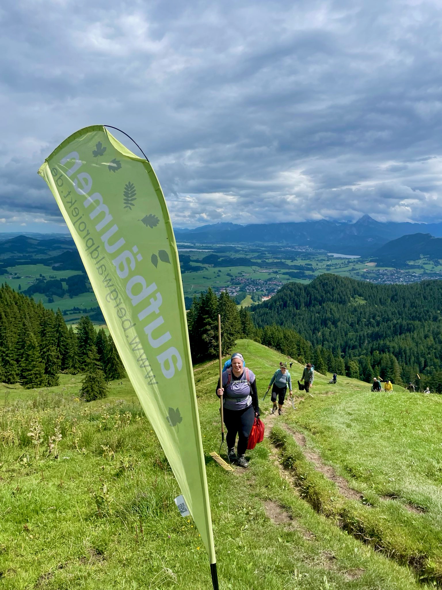 Hinter einer Bergwaldprojekt-Flag kommt eine Gruppe Menschen auf einen Berg hoch gelaufen