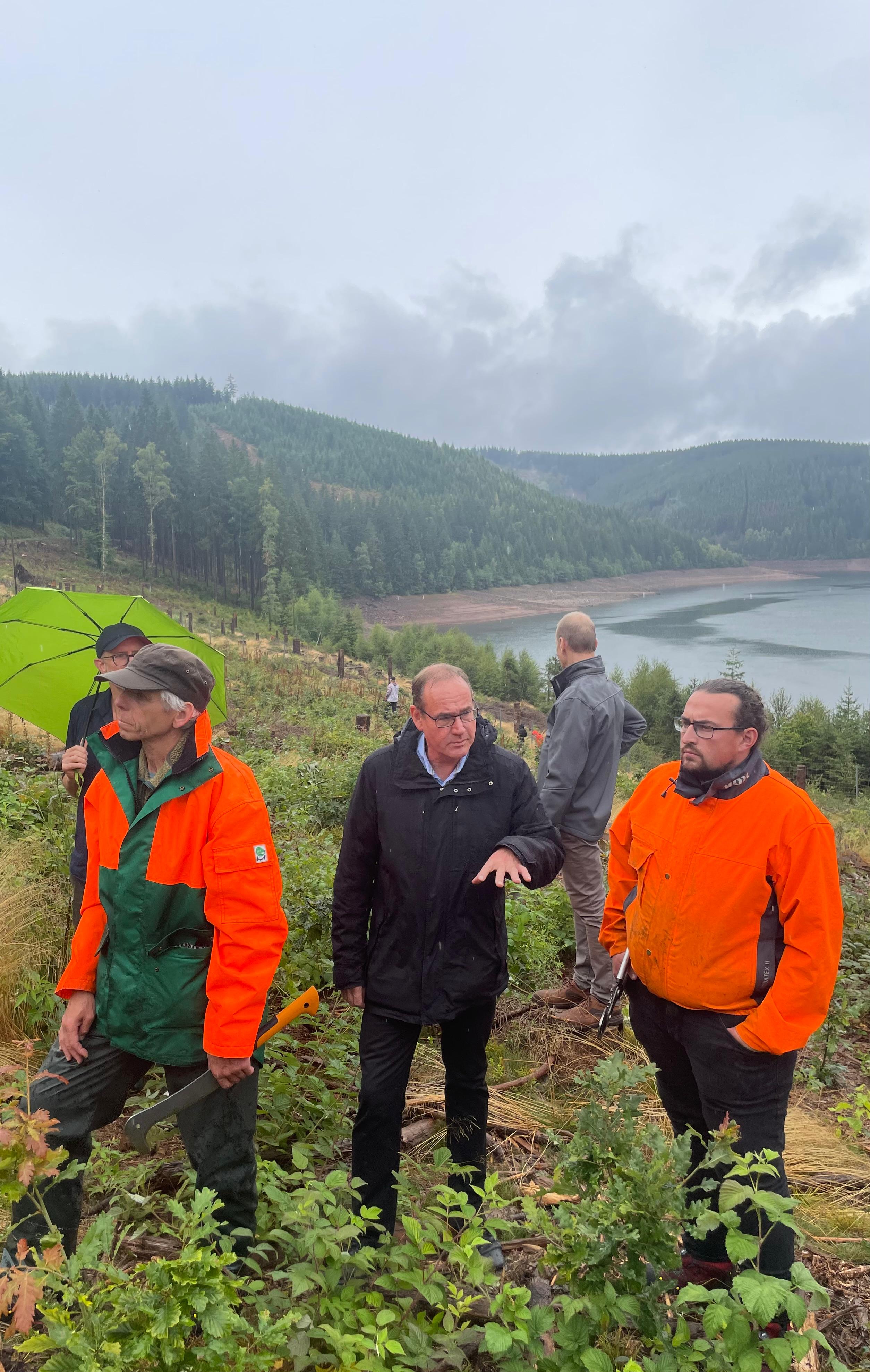 Drei Personen, in der Mitte der thüringische Umweltminister Tilo Kummer, im Hintergrund die Ohratalsperre und arbeitende Freiwillige des Bergwaldprojekts