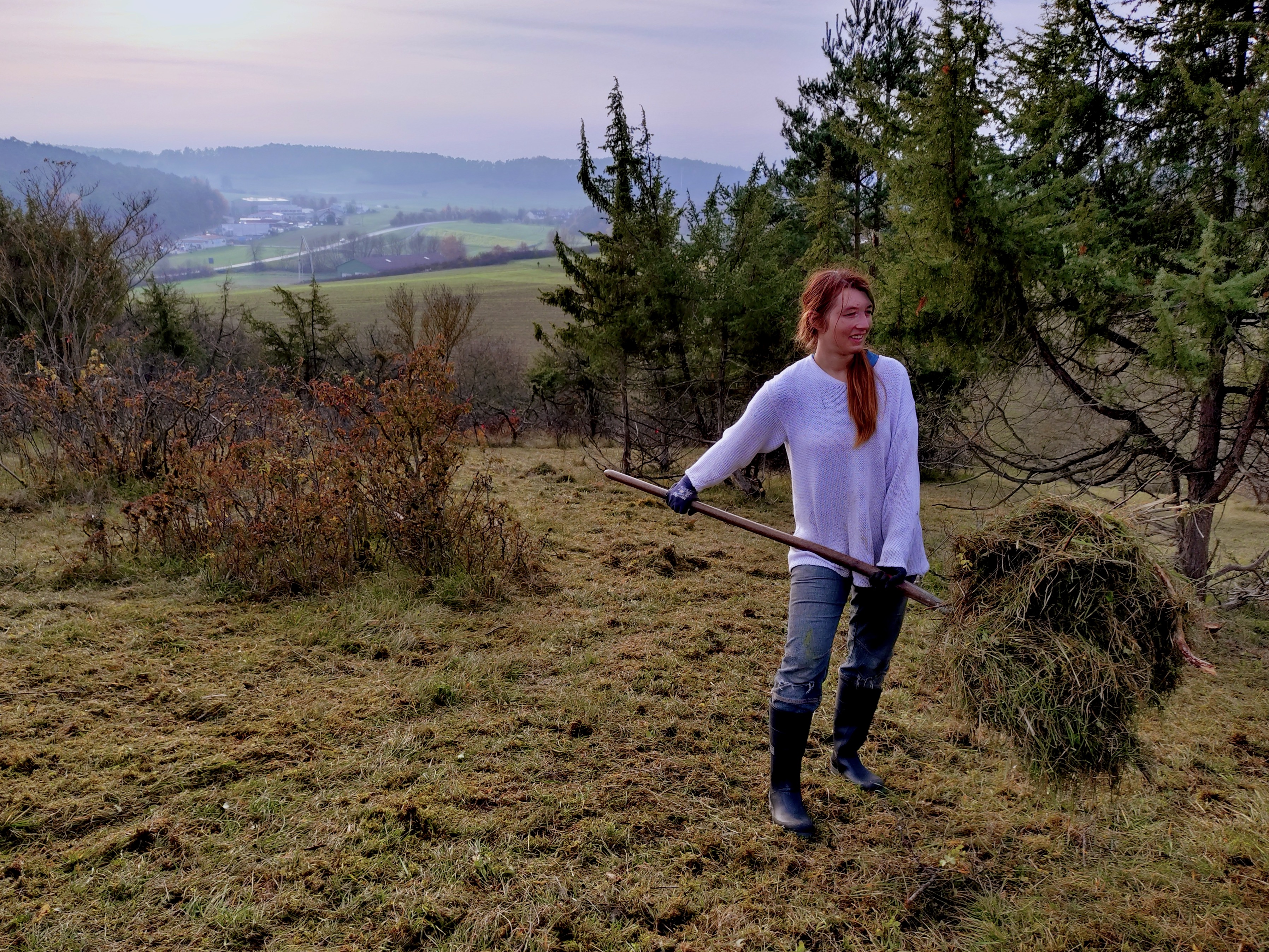 Eine Frau auf einer Rasenfläche mit Wacholder, in der Hand eine Mistgabel mit Grasschnitt darauf