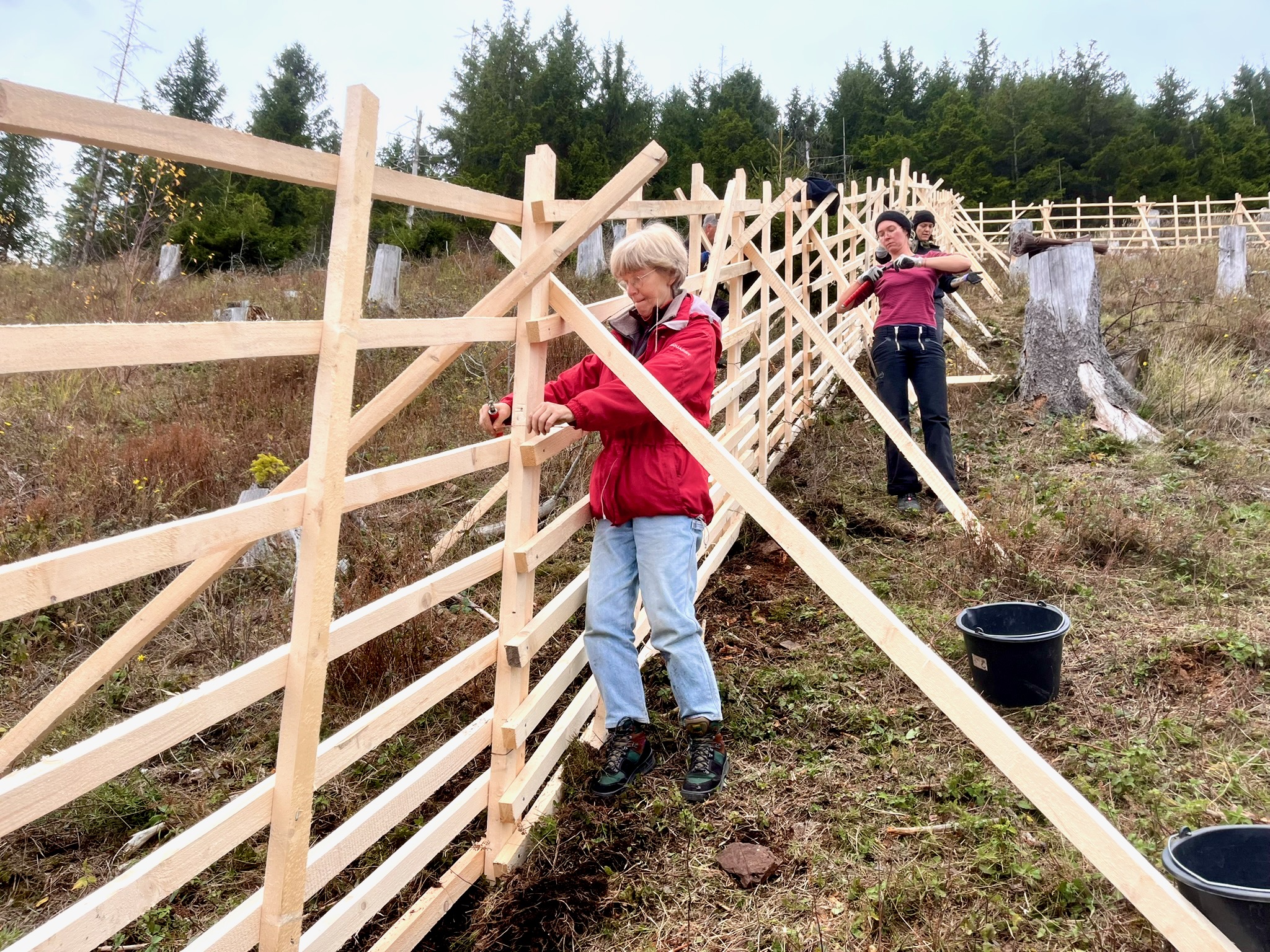 Ein großen Lattenzaun, auf der einen Seite stehen Menschen, die an ihm arbeiten
