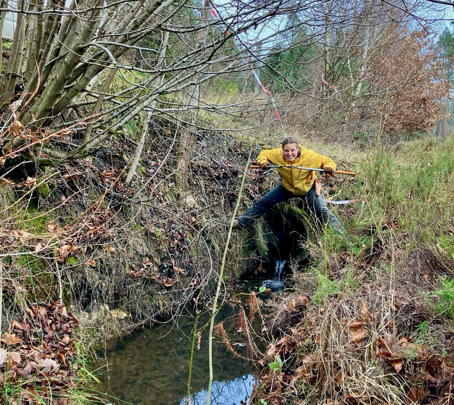Ein Mensch im Friesennerz steht breitbeinig über einem Kanalrohr vor einem Kanal