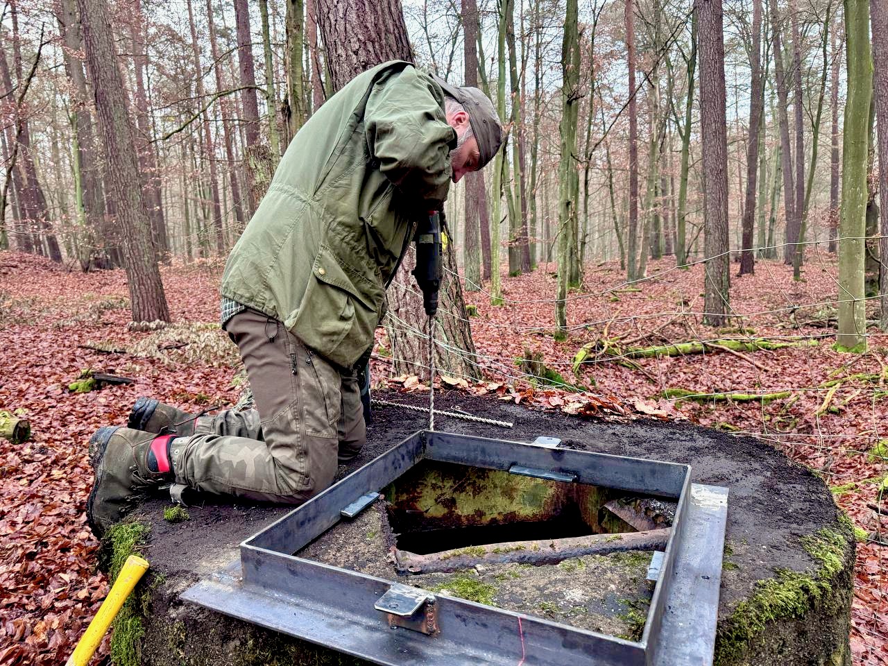 Mann kniet auf rundem Betonklotz im Wald und bohrt mit einem Schlaghammer in den Beton