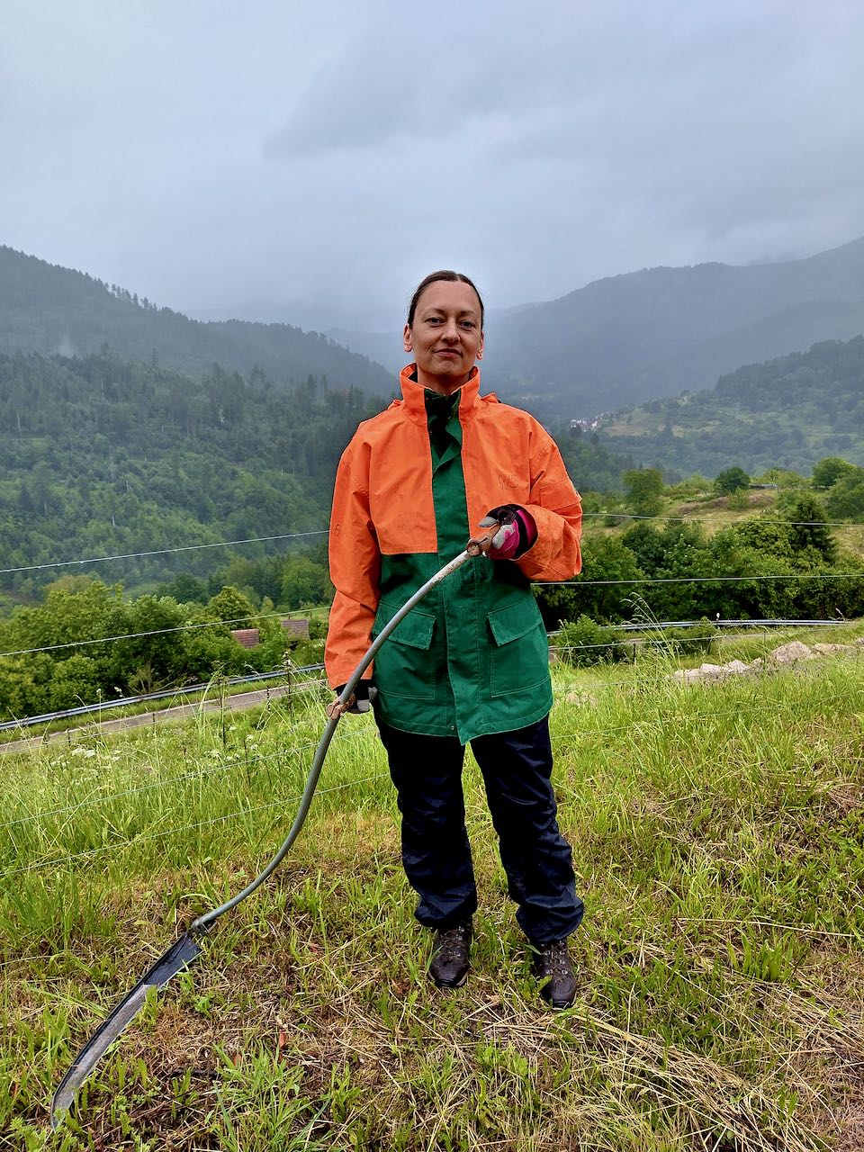 Christina, Freiwillige in Bermersbach Eine Frau steht mit einer Sense auf einer Wiese in einem wolkenverhangenen Tal