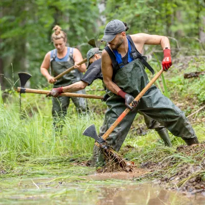 Zwei Teilnehmer arbeiten mit der Projektleiterin mit Wiedehopfhauen in einem Teich