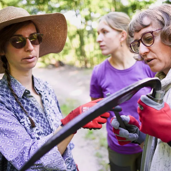 Eine Frau mit großen Strohhut und Sonnenbrille betrachtet mit einer anderen Frau ein Sensenblatt