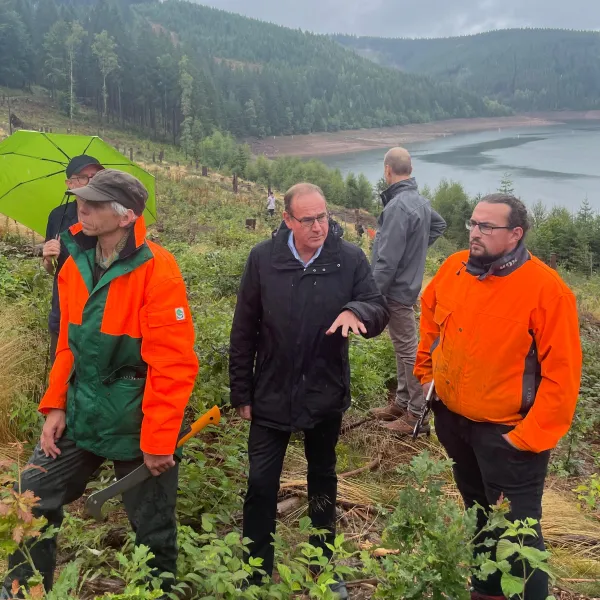 Drei Personen, in der Mitte der thüringische Umweltminister Tilo Kummer, im Hintergrund die Ohratalsperre und arbeitende Freiwillige des Bergwaldprojekts