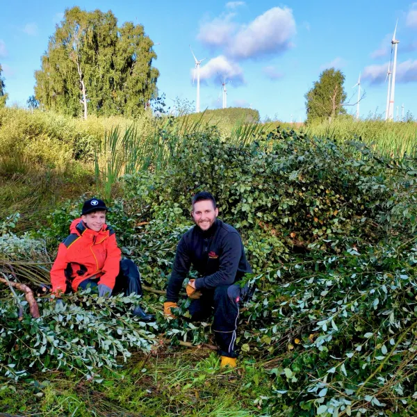 Zwei Menschen knien vor einer Böschung, im Hintergrund Windräder