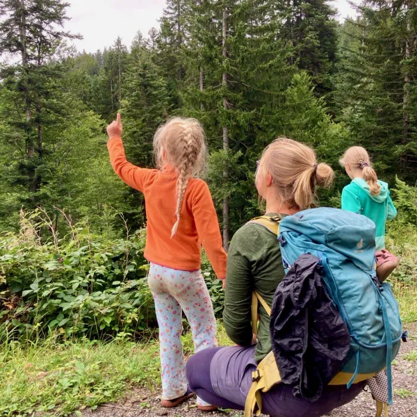 Zwei Kinder stehen mit dem Rücken zum Betrachter, eine Frau kniet zwischen ihnen. Sie schauen in den Bergwald.