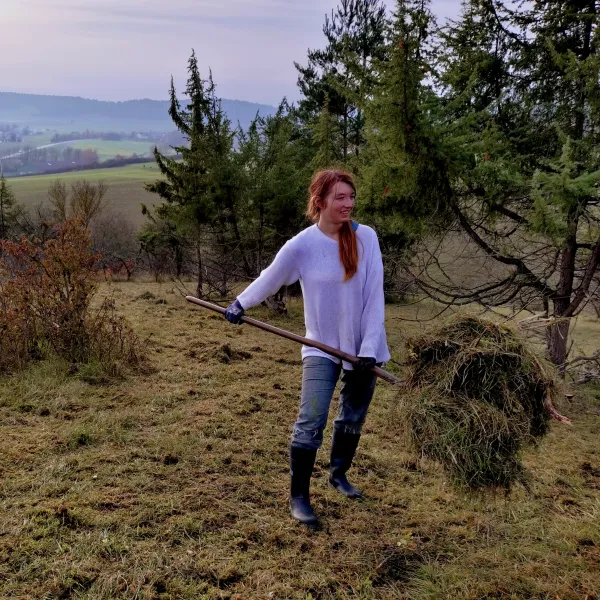 Eine Frau auf einer Rasenfläche mit Wacholder, in der Hand eine Mistgabel mit Grasschnitt darauf