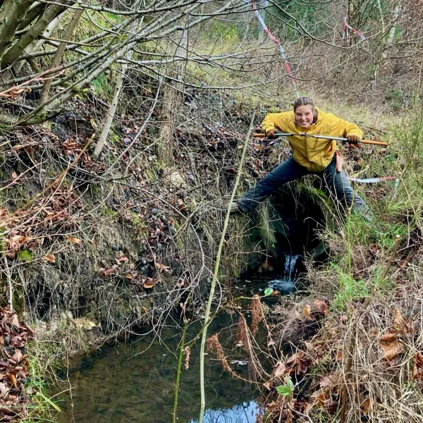 Ein Mensch im Friesennerz steht breitbeinig über einem Kanalrohr vor einem Kanal