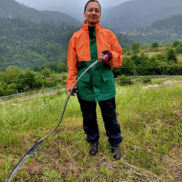 Eine Frau steht mit einer Sense auf einer Wiese in einem wolkenverhangenen Tal