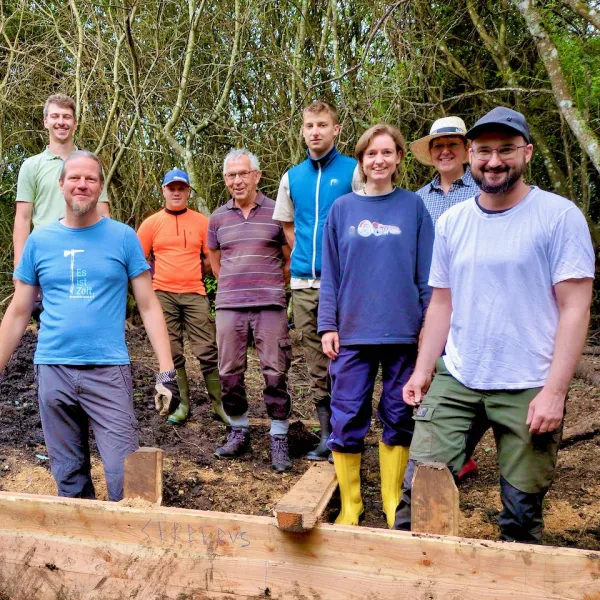 Neun Menschen stehen hinter einer kniehohen Bretterwand vor einer Gehölzgruppe